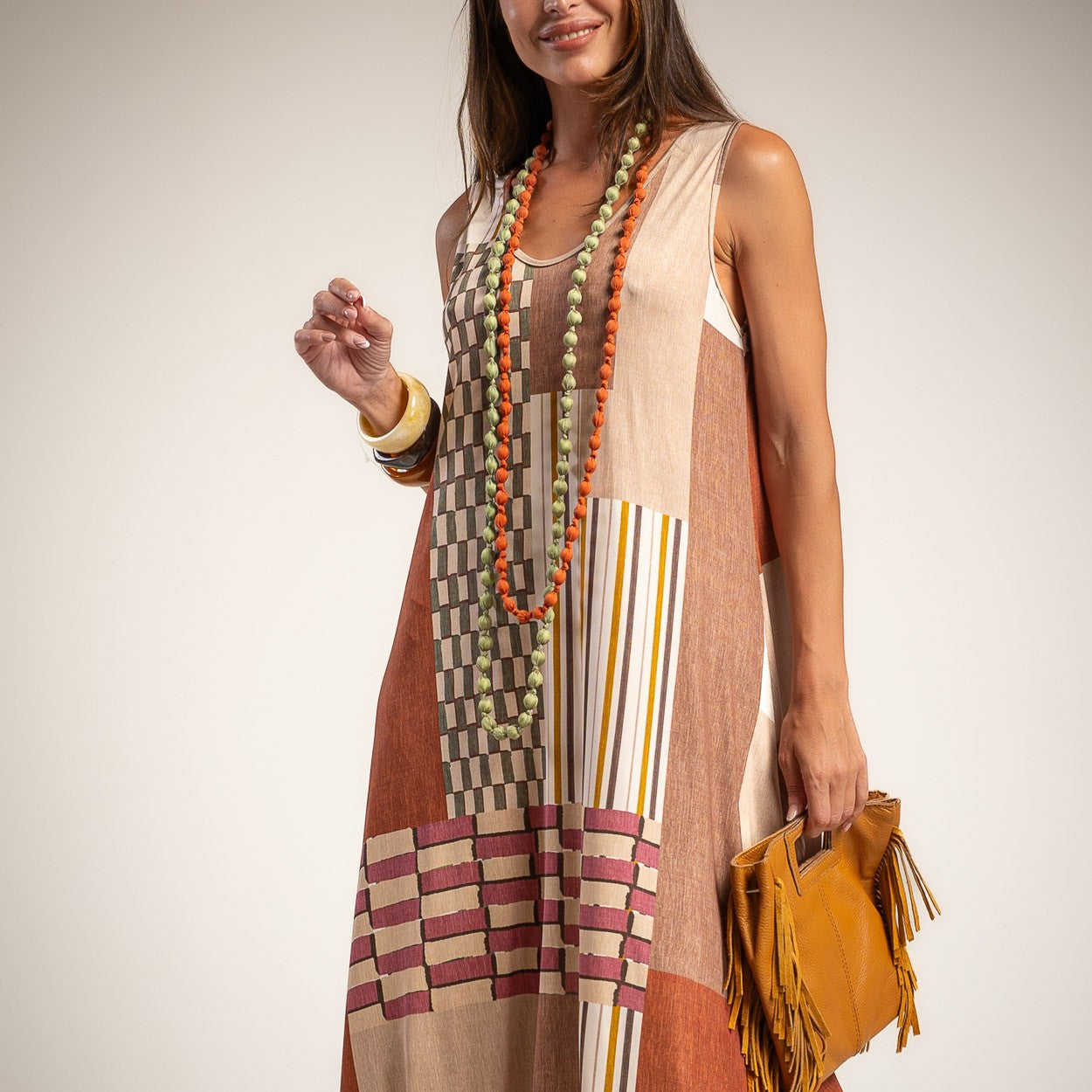 A woman wearing a sleeveless patchwork maxi dress with beige, brown, and rust-orange blocks, striped details, and a geometric pattern near the hem.