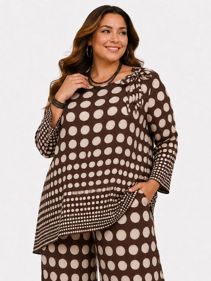 A woman modeling a matching two-piece outfit featuring a brown and beige polka dot pattern, consisting of a long-sleeve ruffled blouse and wide-leg pants, against a plain light gray background.
