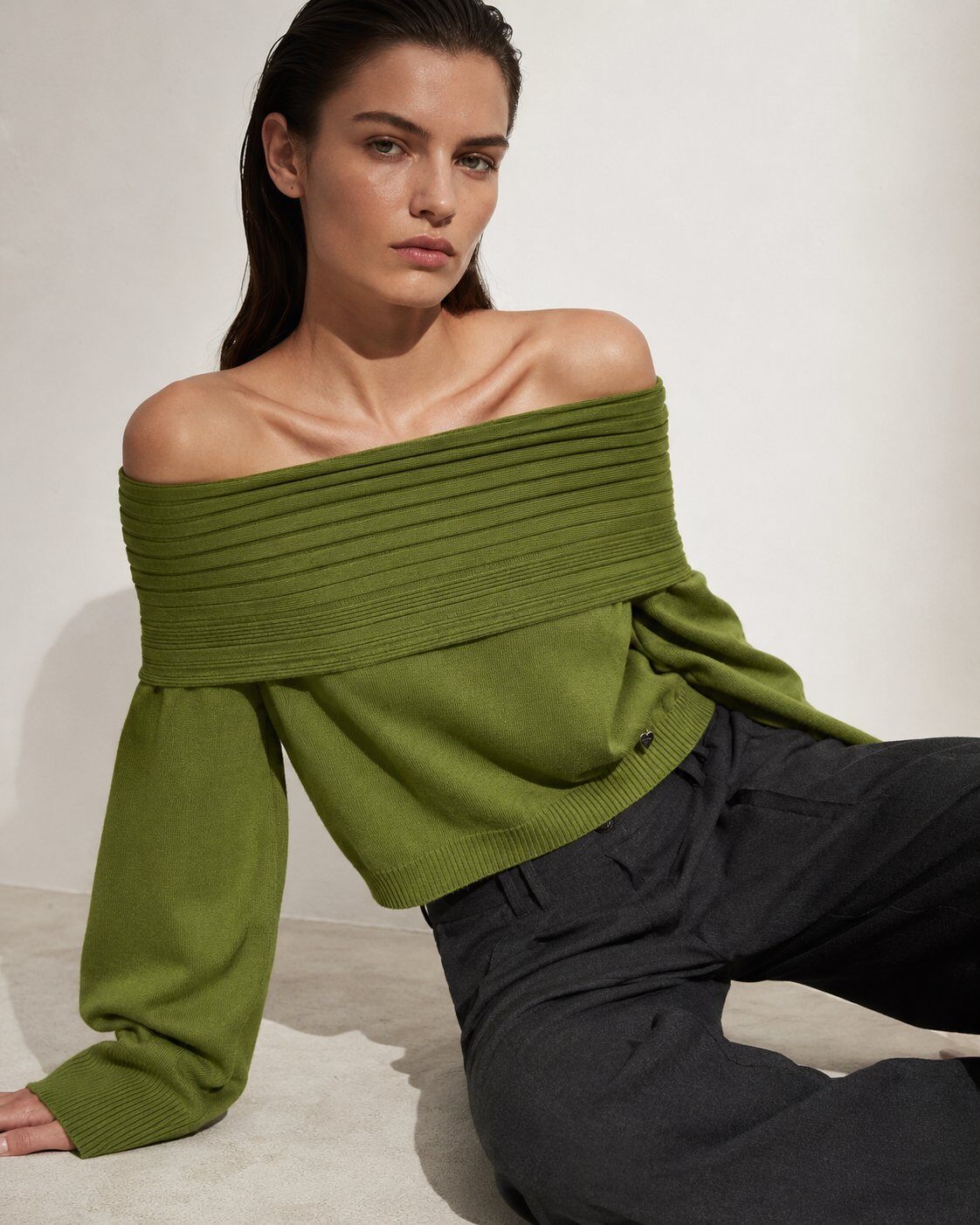 A woman wearing an olive green off-the-shoulder ribbed knit sweater and dark charcoal trousers, posing against a neutral off-white background.