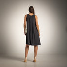 A woman is shown from the back wearing a sleeveless black A-line dress, paired with metallic gold high heels and a small white crossbody bag, set against a studio backdrop with a warm spotlight effect.