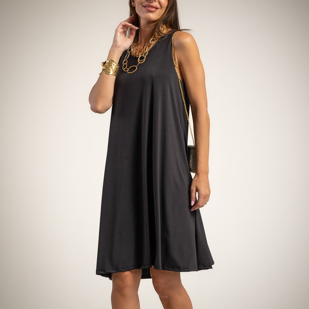 A woman wearing a sleeveless black A-line midi dress with a relaxed fit and side pockets, styled with gold jewelry and a small black crossbody bag, against a light beige studio background.