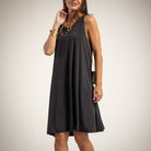 A woman wearing a sleeveless black A-line midi dress with a relaxed fit and side pockets, styled with gold jewelry and a small black crossbody bag, against a light beige studio background.