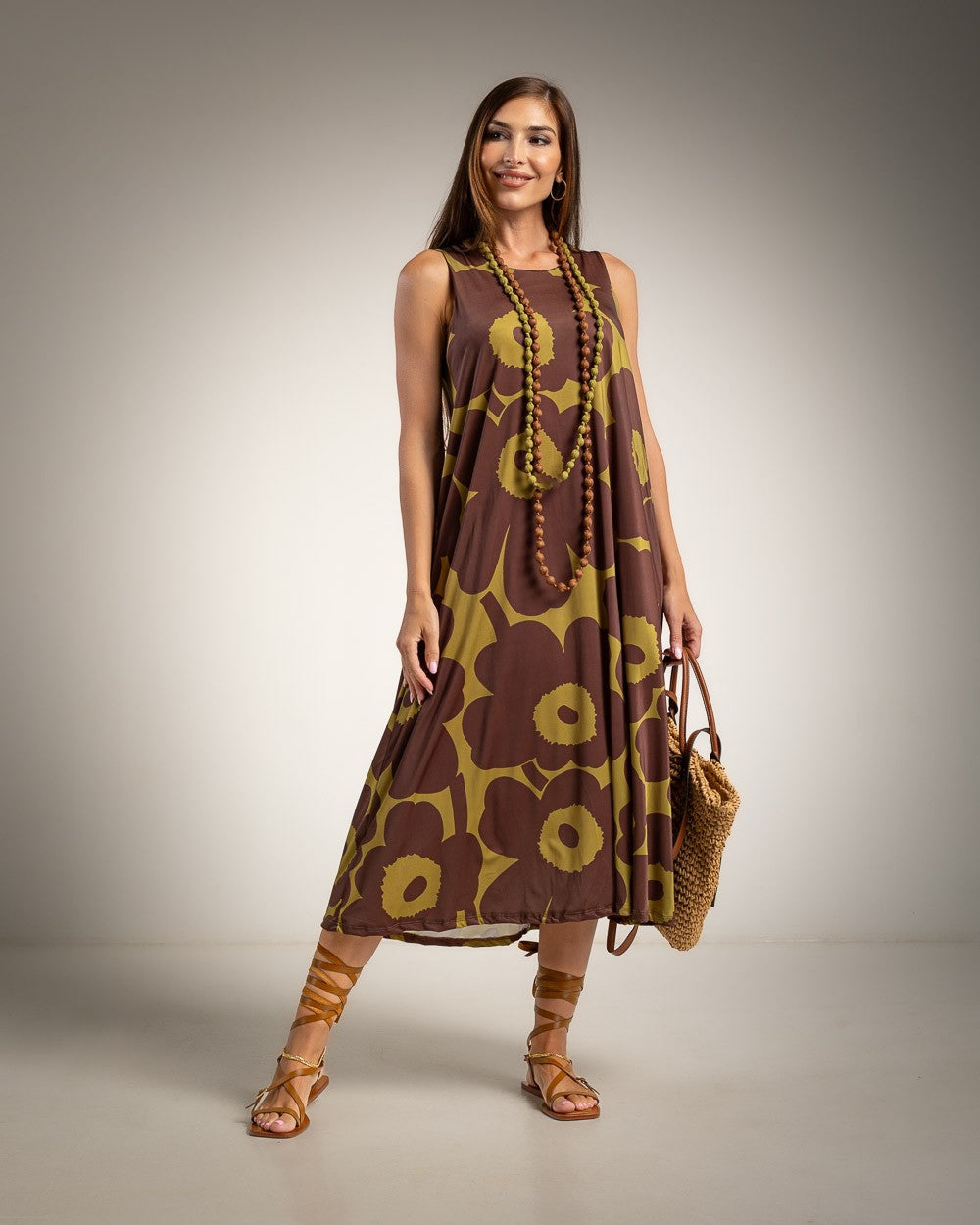 A woman modeling a sleeveless brown and olive green floral midi dress, styled with a long beaded necklace, brown strappy sandals, and a woven straw bag against a neutral studio background.