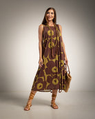 A woman modeling a sleeveless brown and olive green floral midi dress, styled with a long beaded necklace, brown strappy sandals, and a woven straw bag against a neutral studio background.