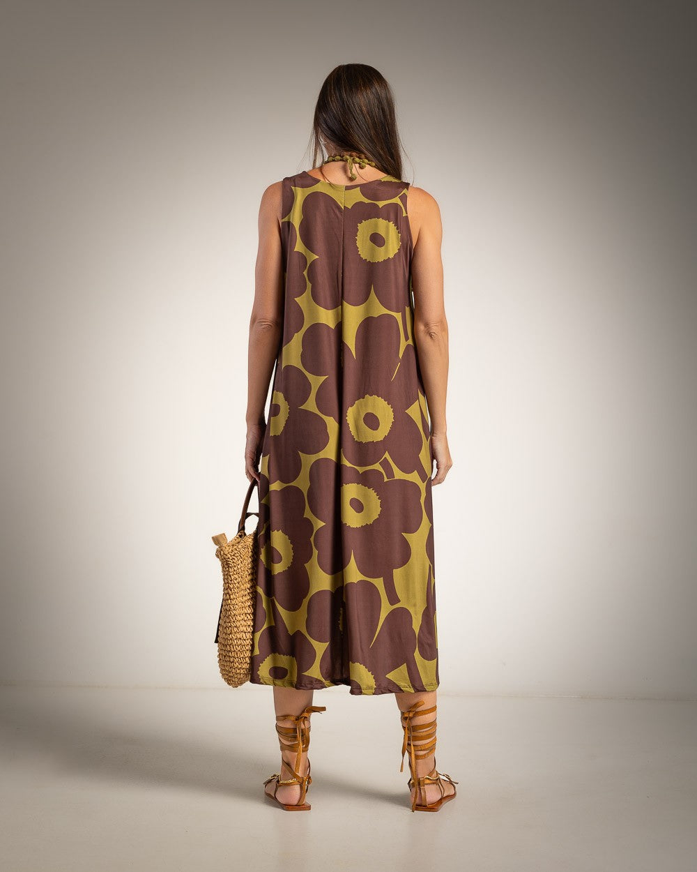 A woman stands with her back to the camera wearing a sleeveless brown dress with a large olive-yellow floral pattern. She is holding a woven straw bag and wearing brown strappy high-heeled sandals against a neutral studio background.