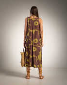 A woman stands with her back to the camera wearing a sleeveless brown dress with a large olive-yellow floral pattern. She is holding a woven straw bag and wearing brown strappy high-heeled sandals against a neutral studio background.