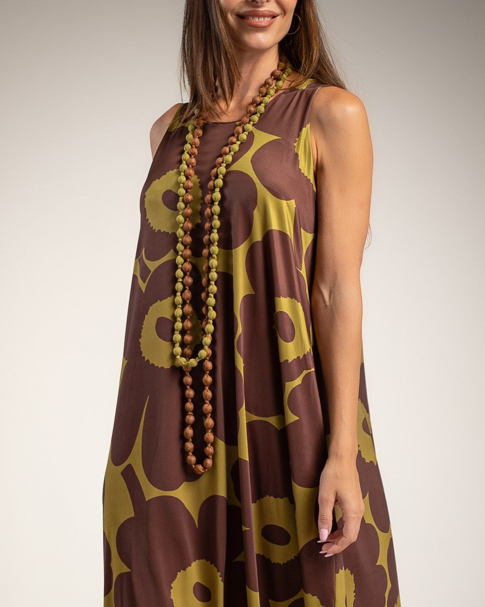 A woman wearing a sleeveless brown and olive yellow floral print dress, accessorized with a long wooden bead necklace, against a neutral studio background.