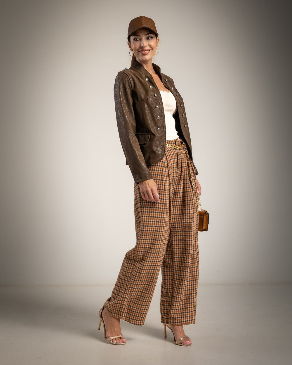 A woman models a brown faux-leather jacket, brown plaid wide-leg trousers, a brown cap, and beige high heels against a neutral studio background.