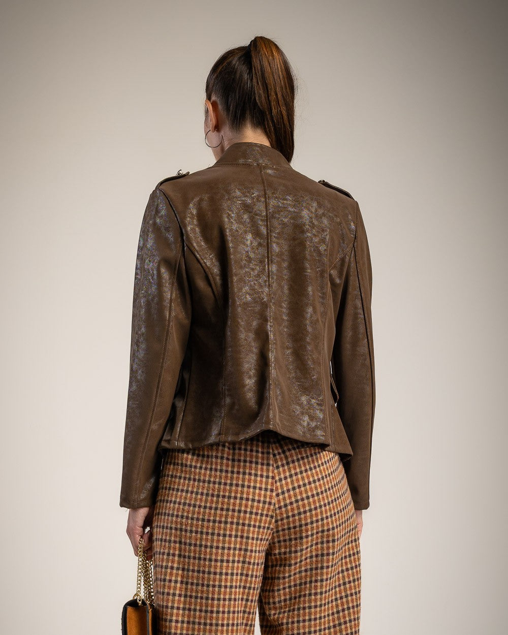 A model shown from the back wearing a brown textured leather jacket and brown-and-orange plaid trousers, holding a small brown handbag against a plain light gray studio background.