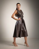 A woman modeling a sleeveless, dark brown faux-leather midi dress with a tie waist, paired with gold bangles and nude high-heeled sandals, set against a neutral studio background.