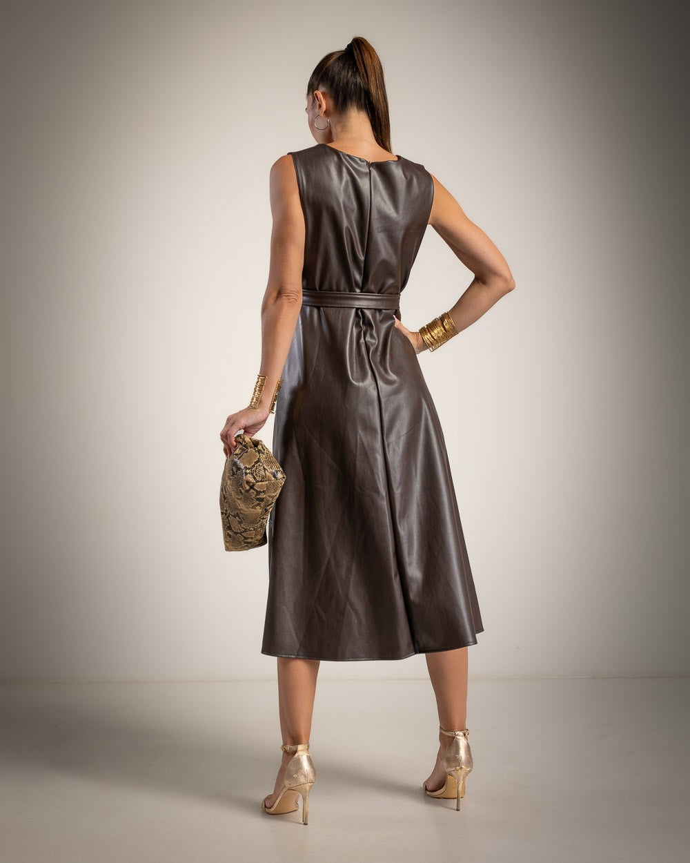 A woman is shown from the back wearing a sleeveless, knee-length brown faux-leather dress with a matching tie belt. She is holding a small patterned clutch and wearing gold high heels and bracelets, set against a neutral studio background.