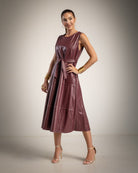 A woman models a sleeveless, tiered midi dress in a deep burgundy faux leather material, styled with gold bracelets, earrings, and nude high-heeled sandals against a neutral studio backdrop.