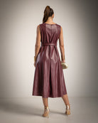 A woman shown from the back wearing a sleeveless, belted midi dress in a glossy burgundy faux leather material, paired with metallic gold high-heeled sandals and holding a small gold clutch against a neutral studio backdrop.