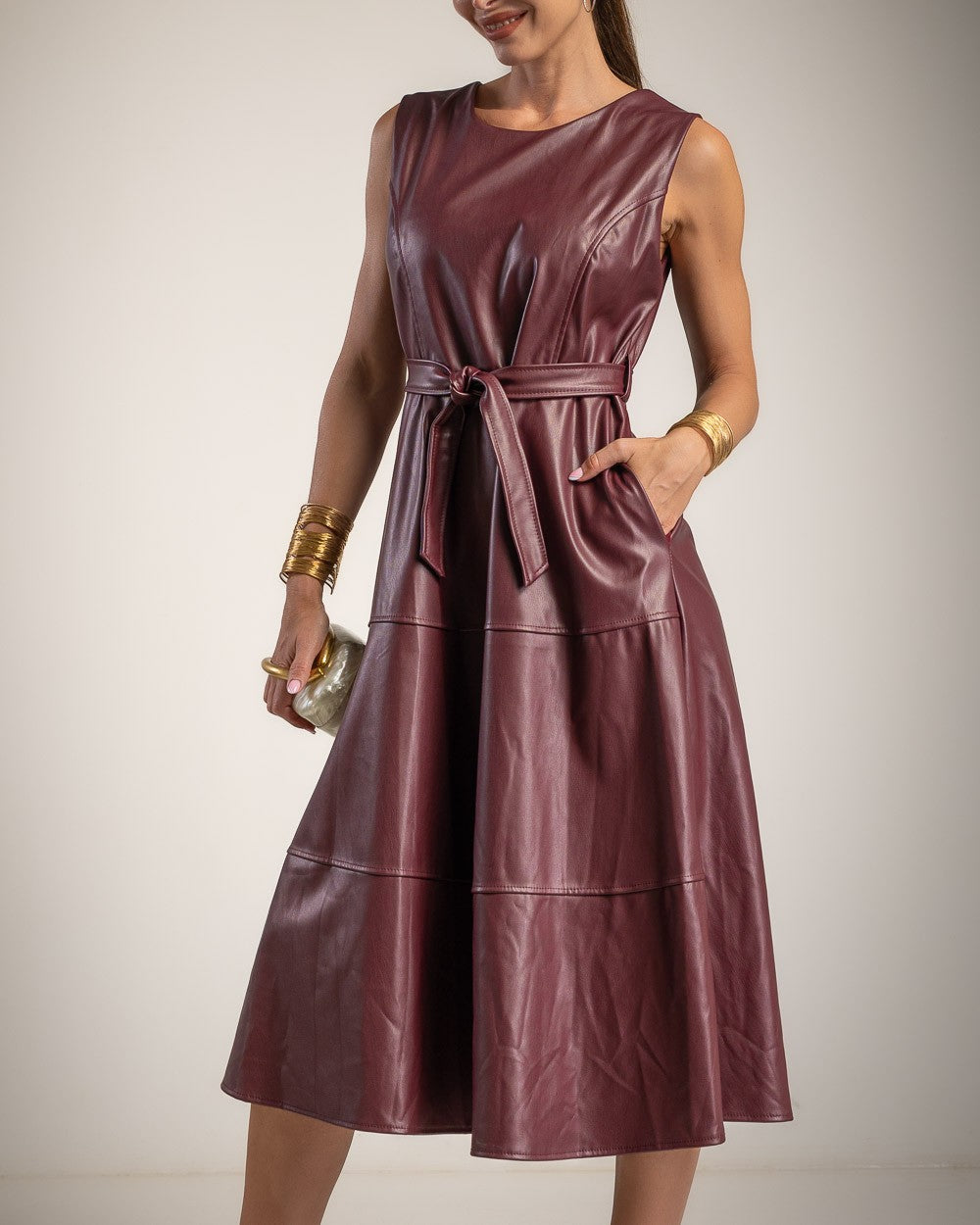 A sleeveless, knee-length faux leather dress in a deep burgundy or plum-brown shade, featuring a tie waist belt and side pockets, modeled against a neutral studio background.