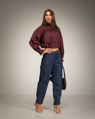 A woman modeling a dark burgundy ruffled long-sleeve crop top and high-waisted dark blue jeans, holding a brown handbag against a neutral gray studio background.