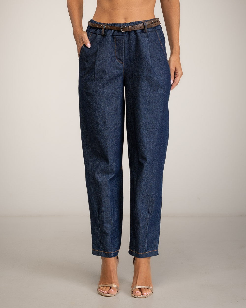 A model is shown from the waist down wearing dark blue denim cropped pants with a brown leather belt, standing against a neutral light gray studio background.