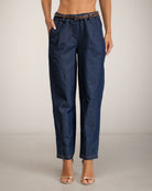 A model is shown from the waist down wearing dark blue denim cropped pants with a brown leather belt, standing against a neutral light gray studio background.
