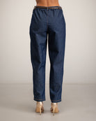 Rear view of a model wearing dark blue denim jeans with a decorative braided waistband and metallic gold high heels, set against a plain light gray studio background.