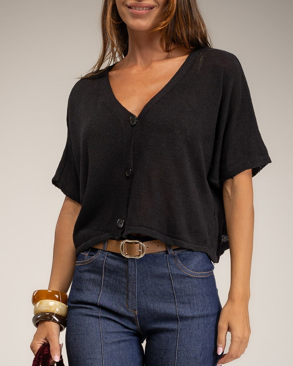 A woman wearing a black short-sleeve V-neck button-front top tucked into blue jeans, photographed against a plain light gray studio background.