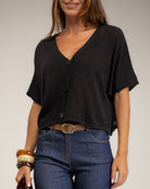 A woman wearing a black short-sleeve V-neck button-front top tucked into blue jeans, photographed against a plain light gray studio background.