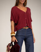 A woman wearing a burgundy short-sleeve button-front top with blue jeans, holding a patterned and velvet handbag against a plain light gray studio background.
