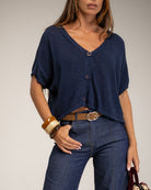 A woman wearing a navy blue short-sleeve button-front top with a V-neck, paired with dark blue jeans and a brown belt, against a plain light gray studio background.