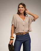 A woman modeling a light beige short-sleeve knit top with a V-neck and button-front detail, paired with dark blue jeans and a brown belt, against a neutral studio background.