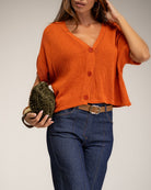 A woman wearing a cropped orange knit cardigan with short dolman sleeves and three front buttons, paired with dark blue jeans and a brown belt, holding a textured green clutch against a light gray studio background.