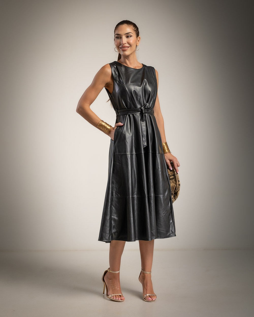 A woman models a sleeveless black faux-leather midi dress with a tie waist, paired with gold bracelets, clear high-heeled sandals, and a patterned clutch, set against a neutral studio backdrop.