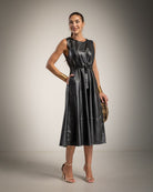 A woman models a sleeveless black faux-leather midi dress with a tie waist, paired with gold bracelets, clear high-heeled sandals, and a patterned clutch, set against a neutral studio backdrop.