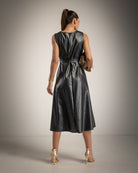 A woman shown from the back wearing a sleeveless black faux-leather midi dress with a matching belt, paired with gold high heels and holding a gold clutch, set against a neutral studio background.