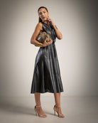 A woman modeling a sleeveless black faux-leather midi dress with a wrap-style waist, holding a textured brown clutch, set against a neutral studio backdrop.