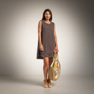 A woman stands in a studio wearing a sleeveless taupe-brown shift dress and holding a woven straw tote bag with colorful floral details.