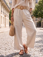 Person wearing a beige knit top and wide-leg pants, holding a woven bag on a cobblestone street.