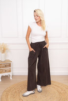 Woman wearing a white top and brown wide-leg pants standing in a room with a wooden side table and decorative items.