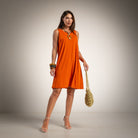 A woman models a sleeveless orange shift dress, holding a gold woven handbag and wearing gold jewelry and clear high-heeled sandals