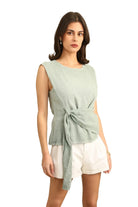Woman wearing a duck green sleeveless top with a tie detail and white shorts on a white background