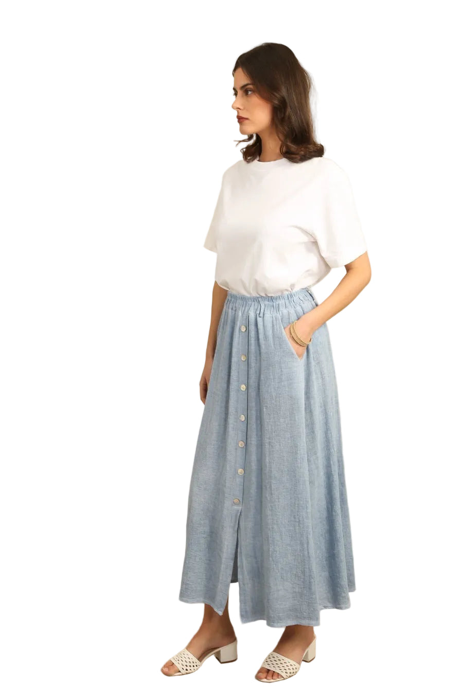Woman wearing a white top and blue skirt with buttons, standing against a white wall.