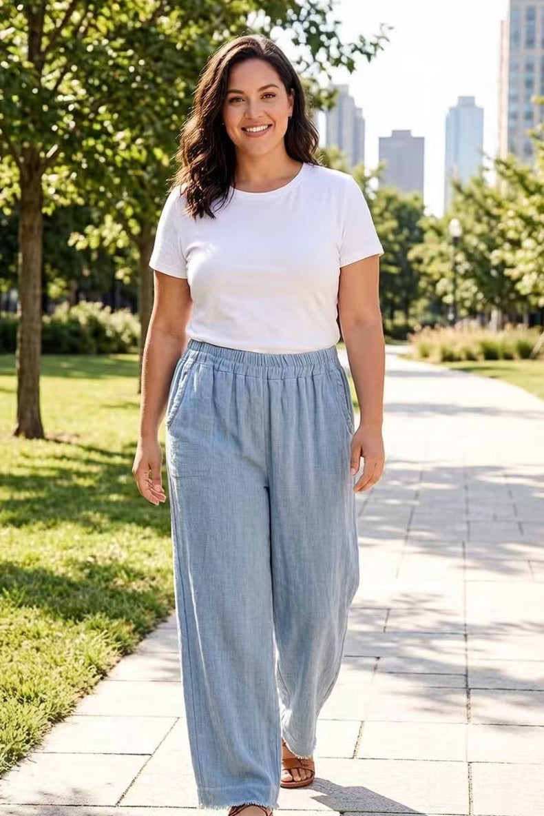 Woman wearing a white t-shirt and light blue pants walking on a sidewalk with trees and buildings in the background.