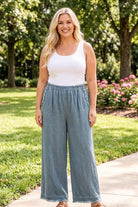 Woman wearing a white tank top and blue wide-leg pants standing on a sidewalk with greenery and flowers in the background.