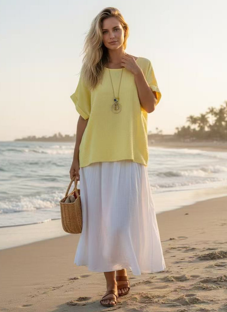 Woman in yellow top and white skirt standing on a beach with ocean and sunset in the background