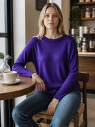 A woman with blonde hair sits on a wooden chair in a cozy cafe setting, wearing a vibrant purple long-sleeve crewneck sweater and blue jeans. A white coffee cup sits on a wooden table next to her.