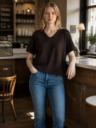 A woman wearing a dark brown short-sleeve V-neck knit top and blue jeans stands in a cafe setting.