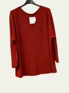 A rust red long-sleeve knit top hanging on a black hanger against a white wall.