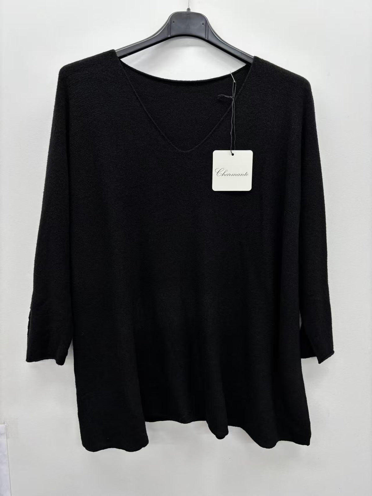 A black long-sleeve knit top hanging on a black hanger against a plain white wall, with a white brand tag attached near the neckline.