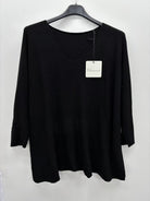 A black long-sleeve knit top hanging on a black hanger against a plain white wall, with a white brand tag attached near the neckline.