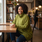 A woman with curly hair sits at a wooden table in a cozy cafe, wearing an oversized olive green turtleneck sweater, blue jeans, and holding a white mug.