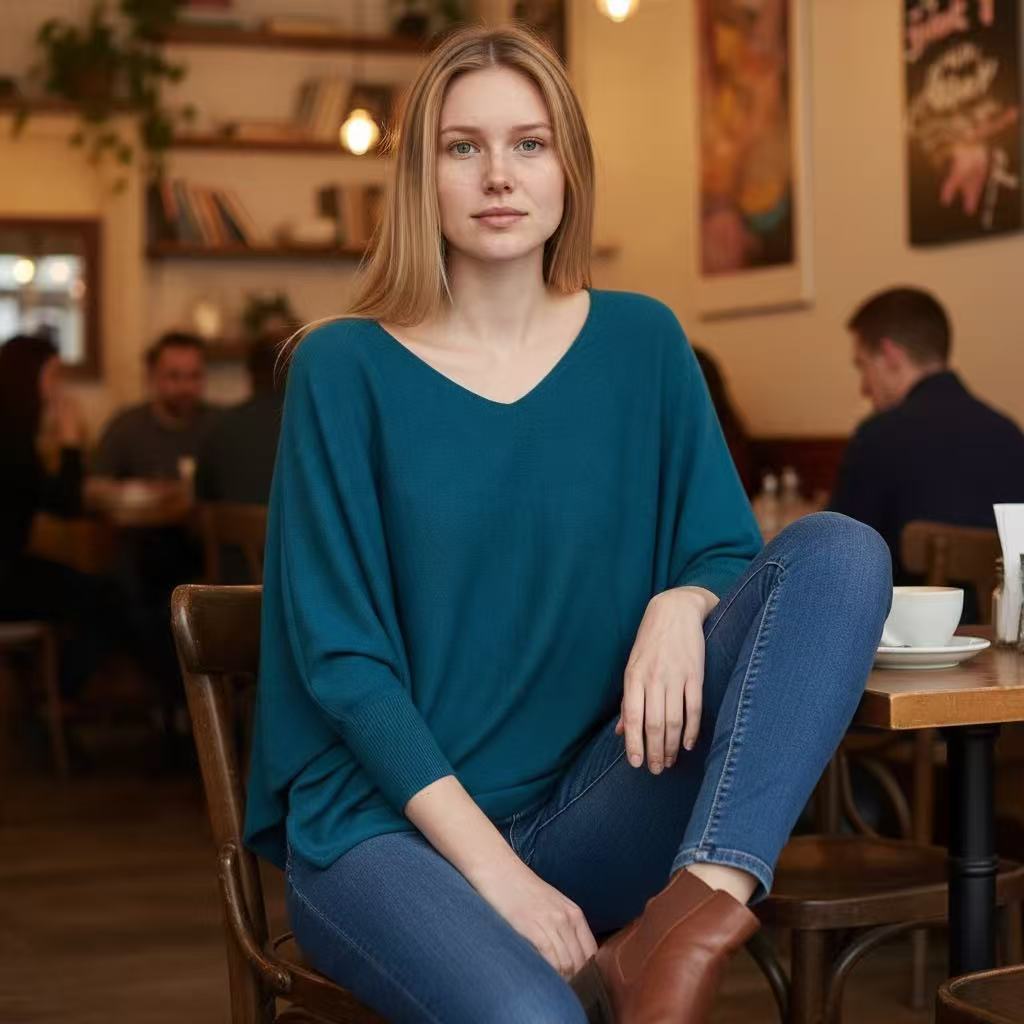 A woman sits in a cafe wearing a teal V-neck long-sleeve sweater, blue jeans, and brown leather boots. The background shows a warm, softly blurred cafe interior with tables, chairs, and other patrons.