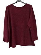 A long-sleeve crewneck knit top in deep burgundy, hanging on a black hanger against a plain off-white wall.