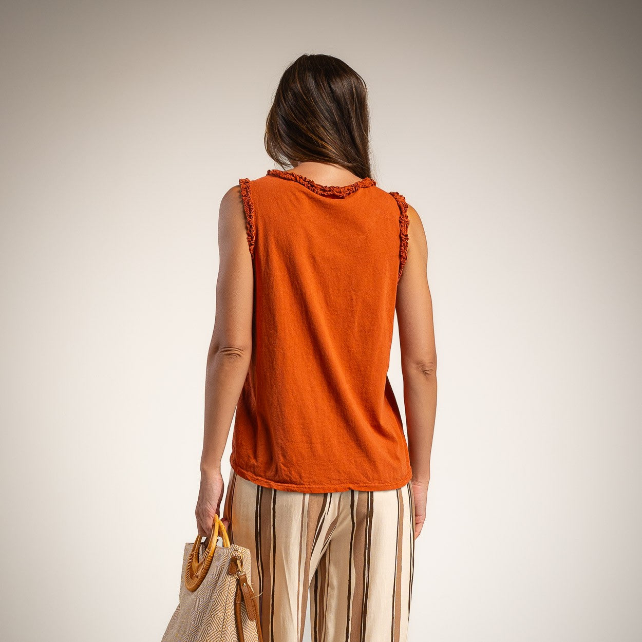 A woman viewed from behind wearing a sleeveless burnt orange top with ruffle trim along the neckline and armholes, paired with beige striped pants and holding a woven straw bag against a plain light gray studio background.